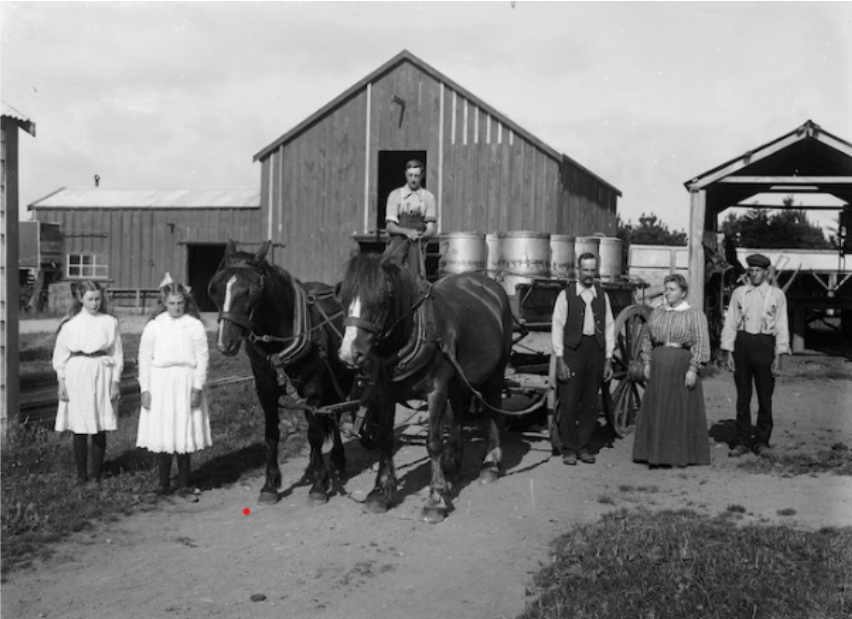 Black and white photograph of a group of people with horses and a cart in front of wooden buildings.